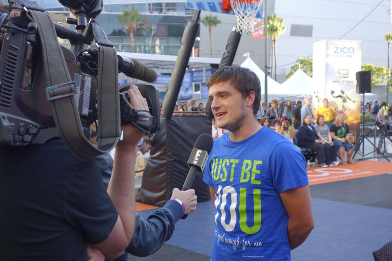 Josh Hutcherson, with long swooping hair, smiles wearing a blue tight-fitting tee that reads "Just Be You" in yellow and white lettering. He stands talking into a microphone held out by a reporter from E! Network and talking into a camera, held by a cameraman whose face is obstructed by the equipment. The interview takes place on a sunny basketball court. Palm trees, a basketball hoop, fans watching sitting on bleachers, and white tents are visible in the background. 