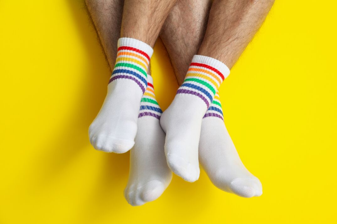 Two men show off their legs in front of a bright yellow background. Their legs start just under the knee and are hairy and muscular. They were white tube socks with rainbow Pride stripes.