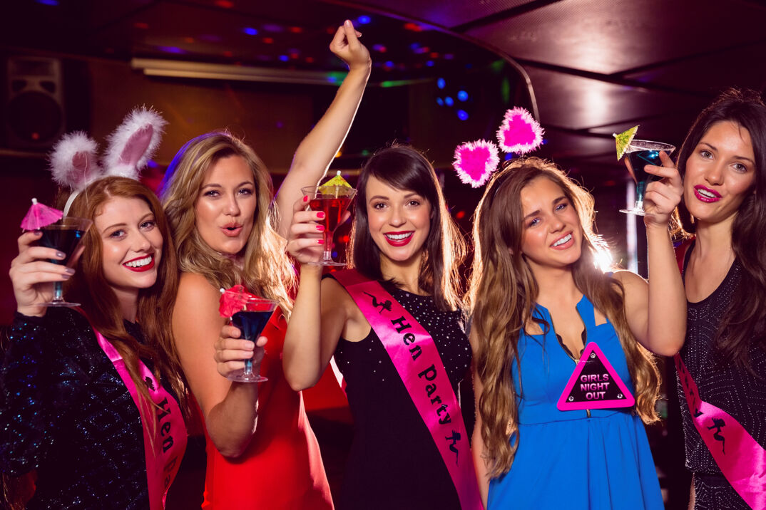 A group of five young woman stand in a nightclub wearing fuzzy bunny ears, pink sashes that read "Hen Party" and smiling. They all hold up fruity cocktails to the camera and wear heavy makeup and fancy party dresses.
