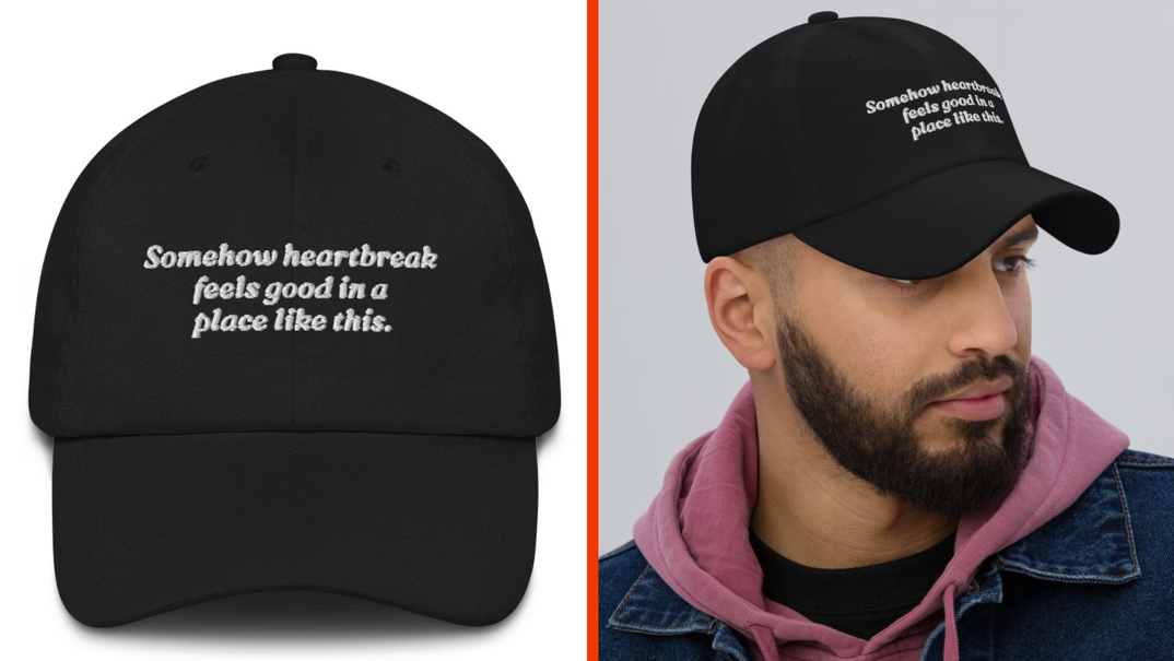 Two-panel image. In the right panel, a man with a thick black beard looks off. He wears a black t-shirt, a purple hoodie, and a dark blue denim jacket. On his head is a black baseball hat that reads "Somehow heartbreak feels good in a place like this" in a white flowy font. In the left panel, a closeup of the baseball hat sitting on a white background.