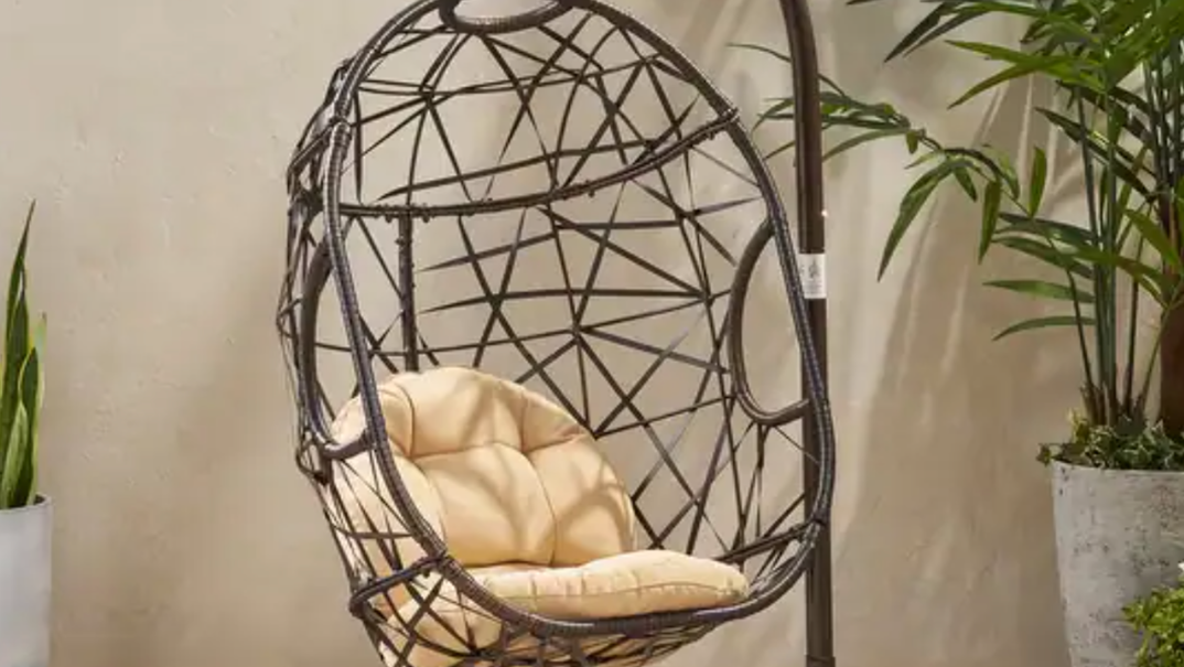 A brown egg-shaped swing chair, with ties connecting across its shape, hangs from a brown stand. In the chair rests two tan cushions. The chair rests in a tan colored room in between two lush potted green plants. 