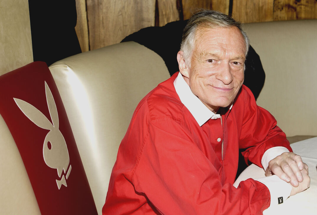 Hugh Hefner, wearing a red button shirt with a white collar, sits on a tan leather couch with the Playboy rabbit logo printed on it. He rests his hands on a white table in front of him. He has gray hair, brown eyes, and smiles softly.