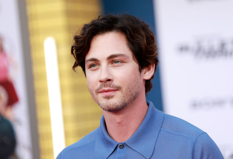 Actor Logan Lerman poses on a red carpet in front of a blurred background. He has long curly brown hair, 5 o'clock shadow, and blue eyes. He smiles softly, wearing a light blue polo shirt buttoned up to his neck.