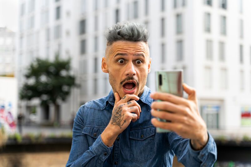 A man with spiky gray dyed hair and the sides of his head shaved stands looking at a smartphone screen shocked in front of a nondescript white building. He has a thin black mustache, tattoos on his left hand which is perched under his chin, and wears a navy blue collared shirt with breast pockets.