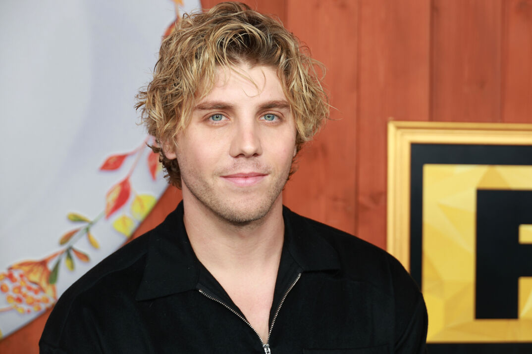 Actor Lukas Gage, with long dirty blonde hair, blue eyes, and blonde stubble, softly smiles in front of a step-and-repeat on the red carpet. He wears a black hoodie, unzipped to expose the top of his smooth chest.