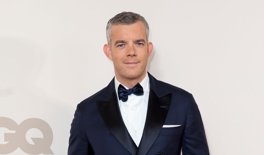 Russell Tovey, with short cropped gray hair and blue eyes, smiles in front of a white step-and-repeat at a GQ event. He wears a dark blue suit jacket over a white buttoned up shirt with a matching bowtie. 