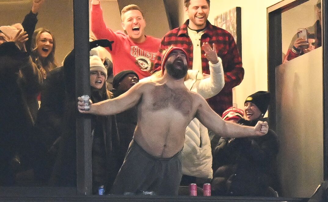 Jason Kelce, holding a beer, sticks his body outside a window at a football stadium in Buffalo, New York. He stands shirtless, showing off his hairy chest, and bellows in excitement. He wears gray sweat pants and is surrounded by cheering Kansas City Chief fans behind him, who remain clothed.