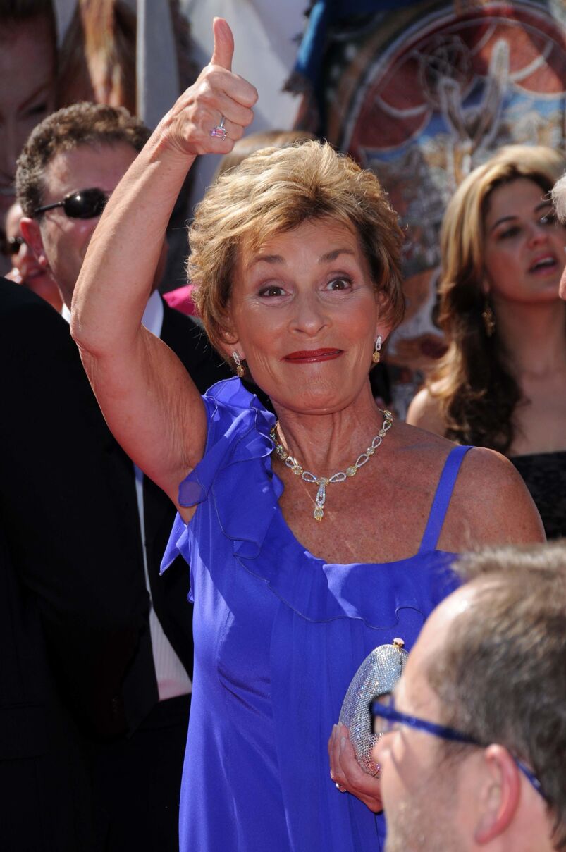 Judge Judy wearing a long blue dress and putting her thumb in the air. 