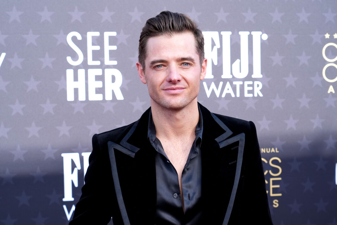 Robbie Rogers at the Critics Choice Awards dressed in a wide black tuxedo and open black dress shirt. 