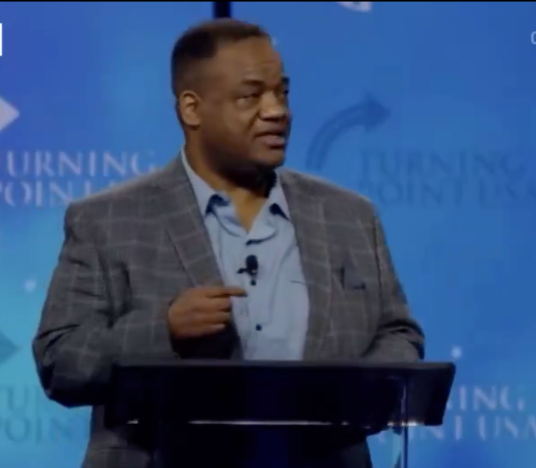 Jason Whitlock wearing a dark sports jacket and blue dress shirt standing behind a lectern on stage. 