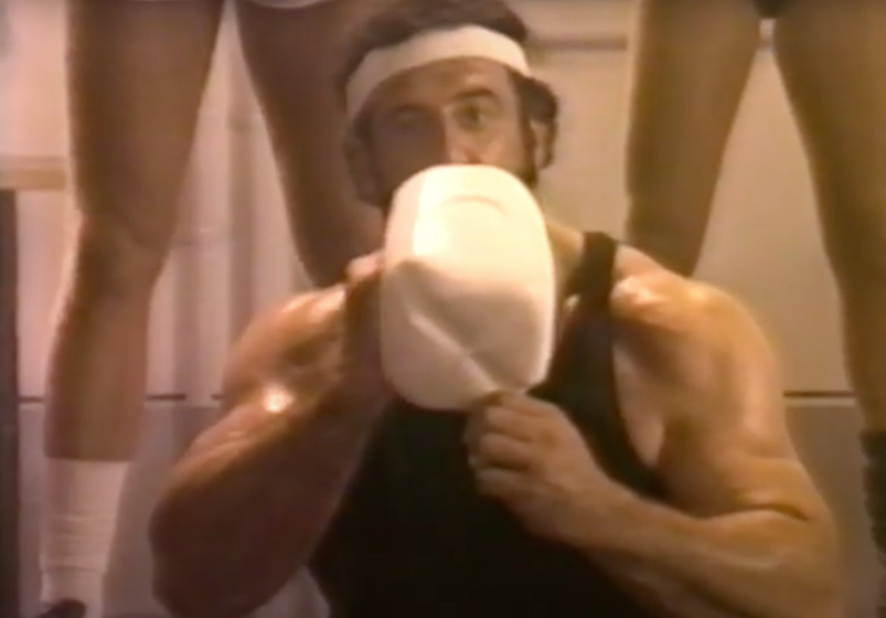 Lyle Alzado drinking a big jug of will while wearing a white headband and right black tank after a workout. 