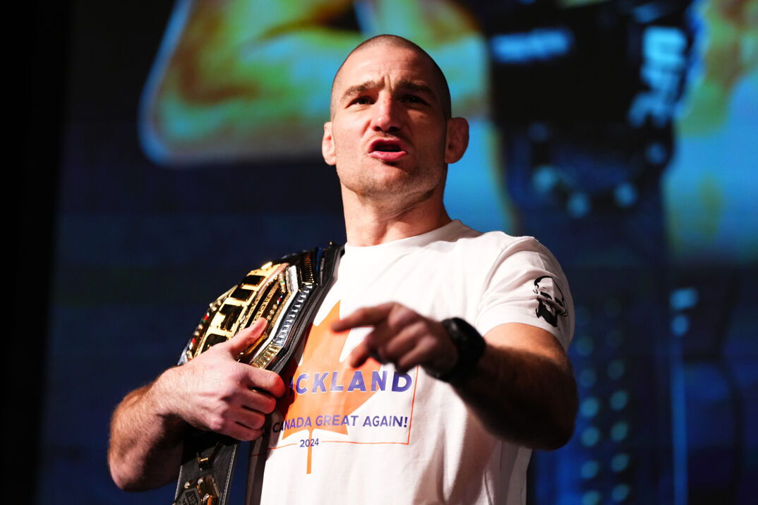 UFC fighter Sean Strickland wearing a white graphic t-shirt with a championship belt over his shoulder. 