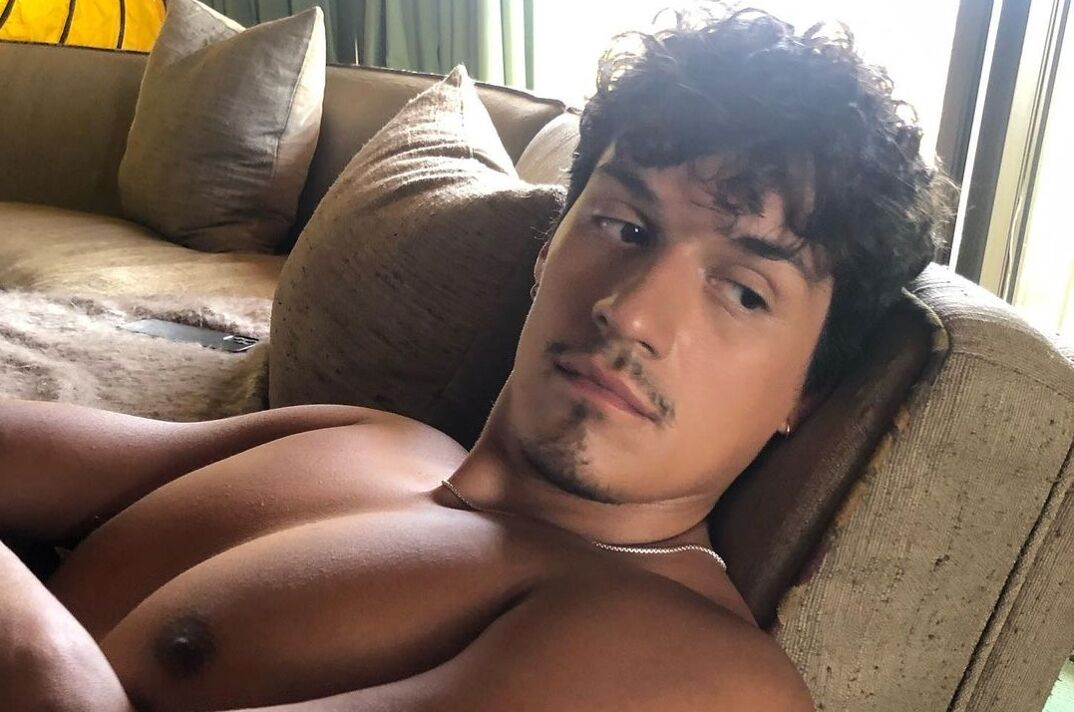 Singer Omar Apollo sits on a brown couch shirtless with his arms crossed. He has curly brown hair and a thin beard and mustache. He wears a silver chain around his neck with his muscular chest and arms on display.