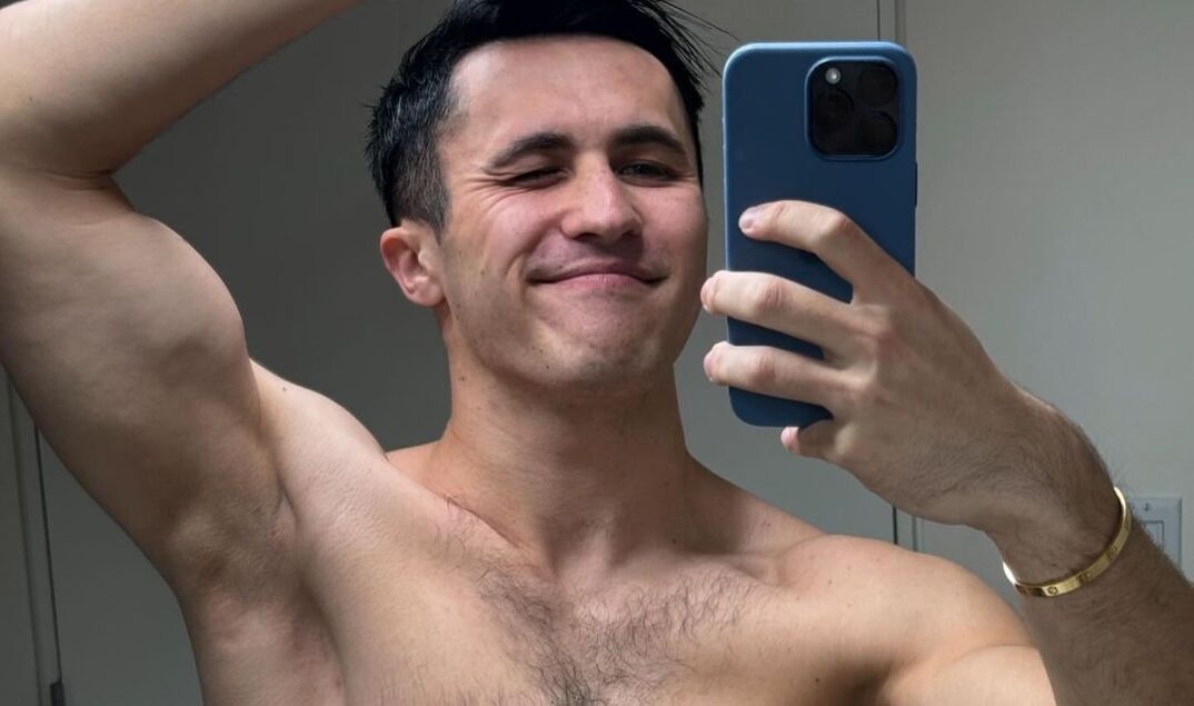 Chris Olsen, with short black hair, winks while taking a selfie on a blue iPhone. He is shirtless and flexing his right bicep, showing off some light chest hair.