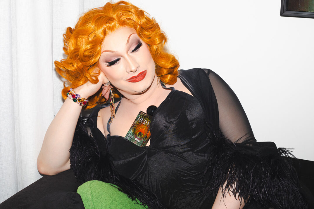 Jinkx Monsoon wears a black negligee and sits resting her head on her hand. In her cleavage is a dazzling orange glass bottle for her perfume 'Hera.' She has long, brilliant, orange curled hair and red lipstick. She sits atop a green couch.