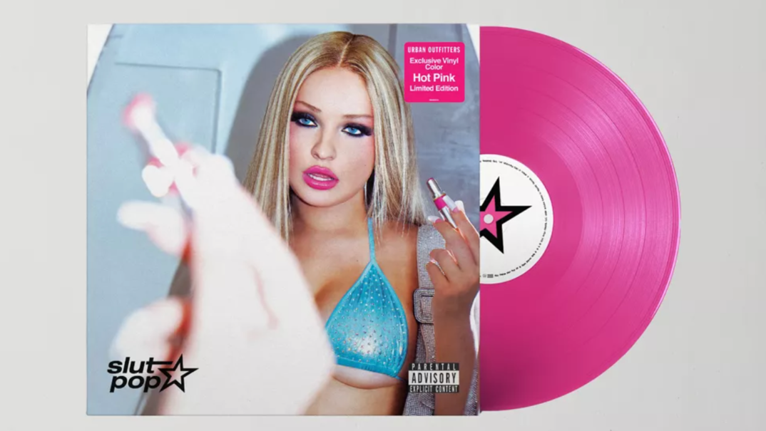 The vinyl album for Kim Petras' 'Slut Pop' is pictured in front of a gray background. A hot pink disc sticks out of its sleeve. On the vinyl cover, Petras touches up her pink lipstick in the mirror while wearing a blue bikini top. In the corner, the words "slut pop" next to a star illustration. In the right corner, a Parental Advisory Explicit Content warning sticker.