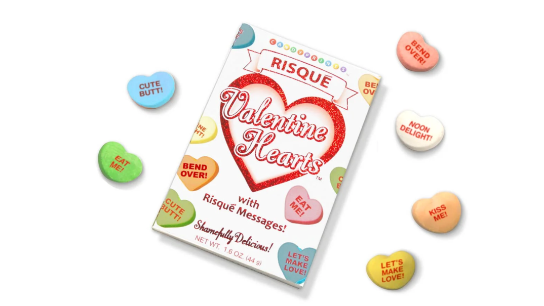 A cardboard carton of heart-shaped candy is pictured laying on a white table. The label reads 'Valentine Hearts with Risqué Messages!' in red text and features illustrations of orange, pink, blue, green, and purple candies with messages on them. Outside of the box, actual candies lay around it reading messages like "Bend Over!", "Kiss Me!", "Cute Butt!", "Eat Me!", and "Noon Delight!" in red text.