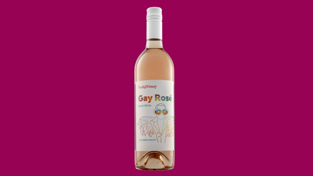 In front of a pink background, a bottle of rosé sits. It has a white cap and a white label reading "Gay Rosé" in rainbow letters over a rainbow line illustration of Elton John in thick glasses.