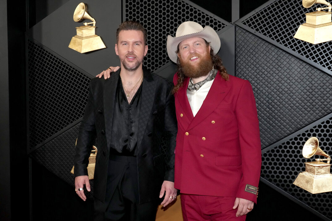 TJ Osborne (with short spiky dirty blonde hair and a beard) stands in a black textured suit, open to reveal a silky black low cut vest revealing some light chest hair. Next to him stands his brother John Osborne, in a white cowboy hat, red suit, and with a bandana around his neck. John has a thick red beard. The two are posed in front of a black step-and-repeat at the Grammys.