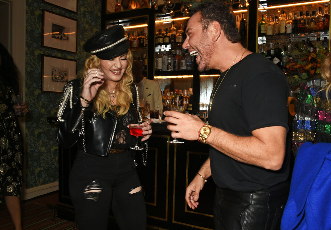 Madonna, wearing a studded black leather outfit and cap with her long curly blonde hair down, stands laughing hold a wine glass filled with a red liquid. Laughing alongside her is Mert Alas in black leather pants, a black tight t-shirt, and a golden necklace and watch. He has brown hair and a thin brown beard. They stand in a dim and crowded New York dive bar.