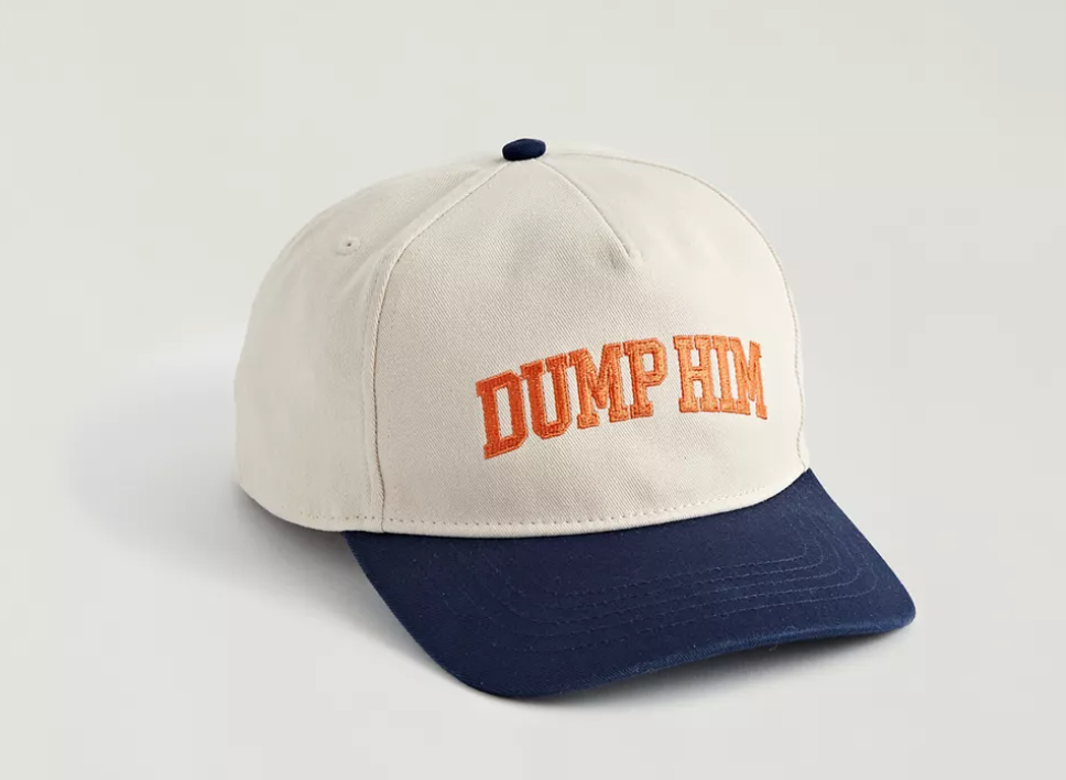 In front of a gray background, a white baseball hat with a blue brim sits. Embroidered across the front of the cap in orange varsity letters: "DUMP HIM."