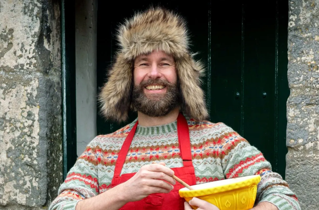 Coinneach MacLeod, who posts on TikTok as the Hebridean Baker, stands in a rustic brick doorway. He wears a furry winter hat covering both his ears and smiles brightly. He has a thick brown beard with hints of gray and stands in a red apron, wearing a colorful warm knit sweater underneath and holding a yellow bowl and wood mixing spoon. 