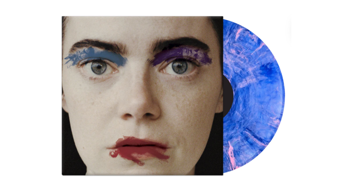 The vinyl for the Poor Things soundtrack is pictured on a white background. Its cover features Emma Stone in character from the film with blue paint smudged along her eyebrows and red paint on her lips. She stares plain faced. A purple and pink paint-splattered vinyl disc peeks out from the sleeve.