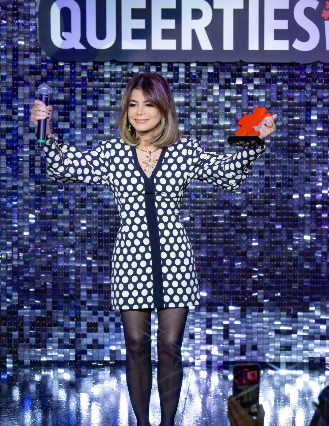 Paula Abdul holds up her arms and smiles while accepting an award at the 2024 Queerties Awards. She wears a black and white polka dot dress and black tights.