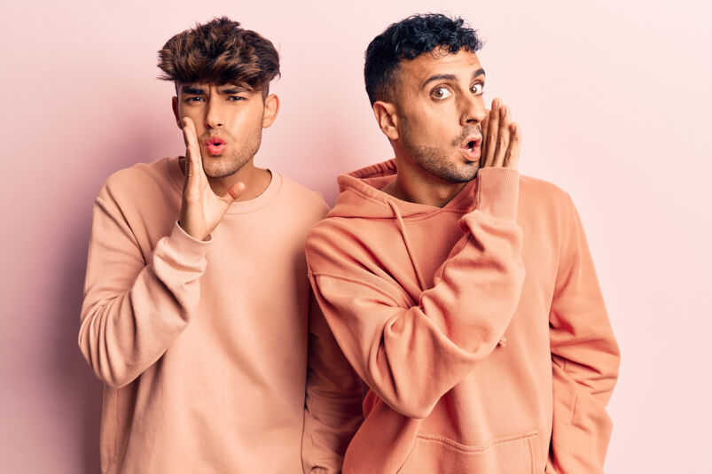 two queer men wearing peach hoodies speaking to each other using gay slang terms. their hands are raised to their mouths as if they're gossiping to each other