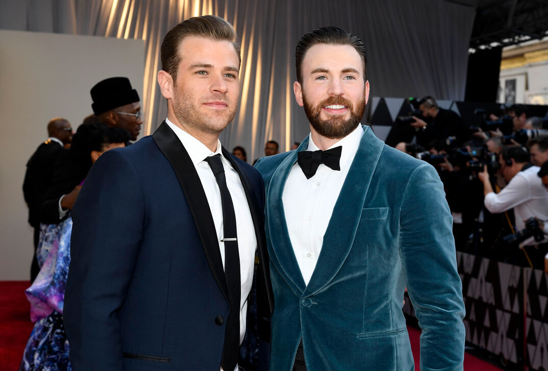 Scott Evans and Chris Evans pose on the red carpet. Scott, on the left, is a hair taller and wears a well-fitting navy tux. He has light blonde hair and five o'clock shadow. His brother, to his right, has strawberry blonde hair and a thick beard and mustache. He wears a turquoise tux.