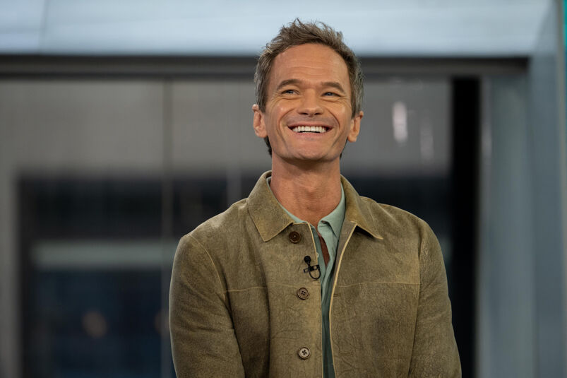 Neil Patrick Harris smiles looking off while sitting at an event. He has short blonde spiked hair and wears a green collared shirt under a darker green overcoat. A small black microphone is clipped to his jacket.