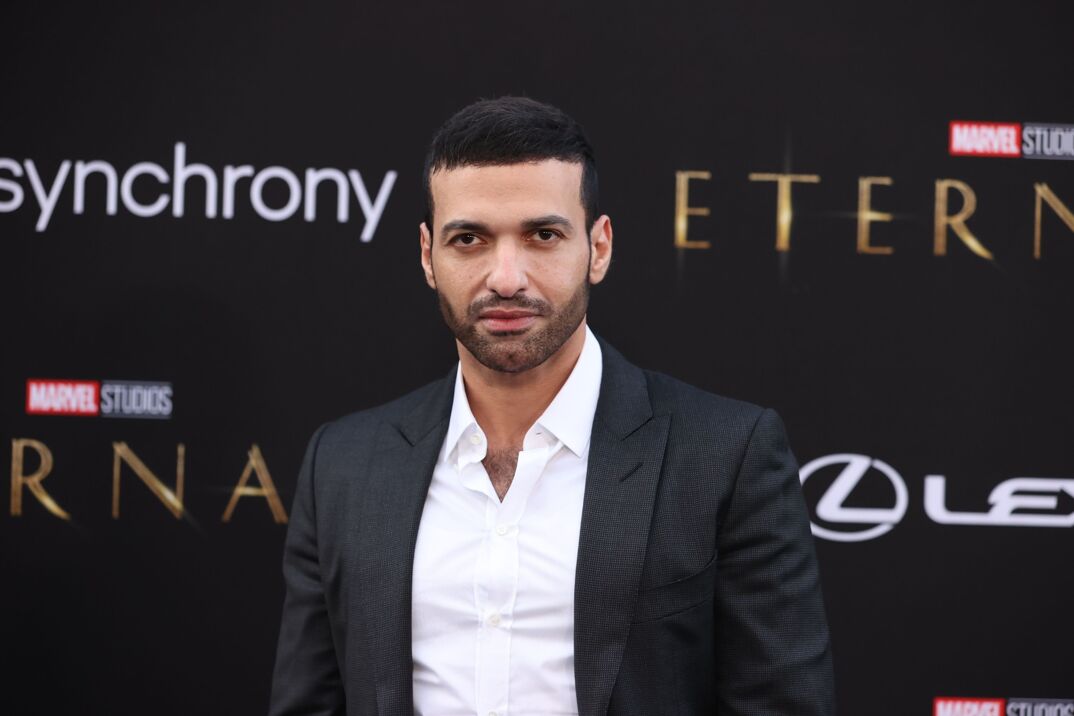 Haaz Sleiman smiles softly in front of a black step-and-repeat. He has buzzed dark hair, dark brows, and a black beard and mustache. He wears a white dress shirt unbuttoned to reveal some chest hair under a black suit jacket.
