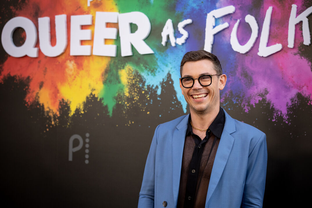 Ryan O'Connell, who has short brown hair and thick rimmed glasses, smiles widely on a red carpet in front of a rainbow-colored sign for "Queer as Folk." He wears a gold chained necklace, and a black sheer button-down shirt under a light blue suit jacket.