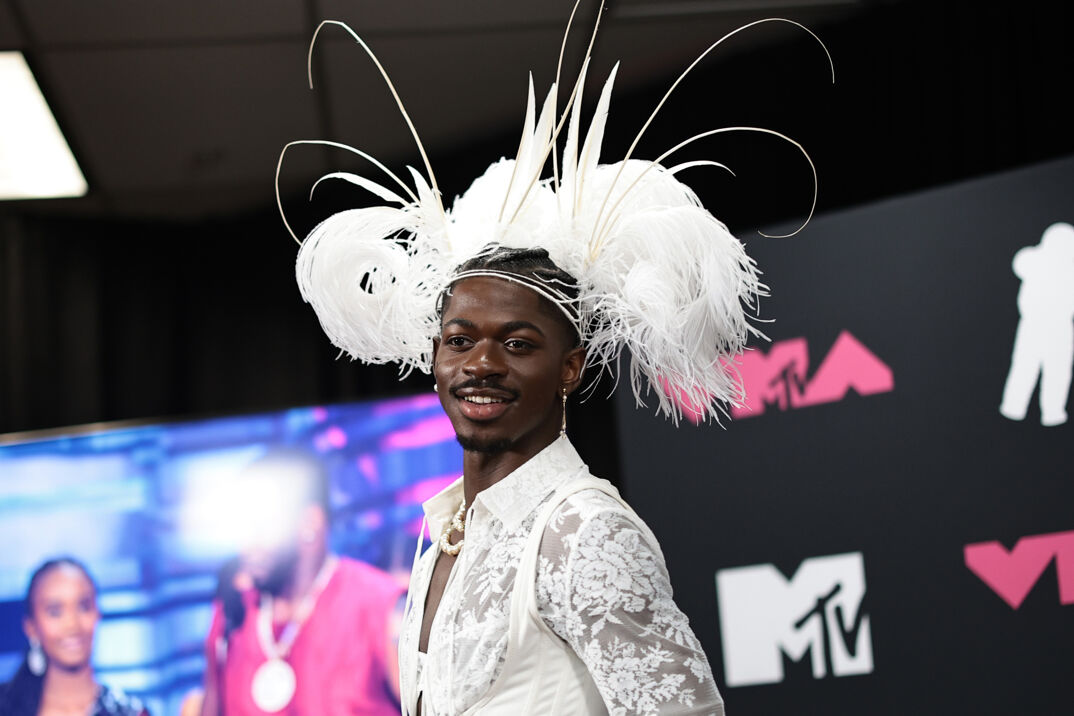 Lil Nas X poses on a red carpet. He wears a white headdress with feathers extending into the air, a pearl necklace, and silver dangling earrings. His dark hair is braided and tucked over his head and the top of his outfit, a sheer and floral collared shirt, is barely visible.