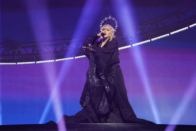 Madonna stands atop of a circular platform singing into a microphone during a Celebration Tour performance. She wears a long, thick black dress with low hanging sleeves and a circular metallic headdress. The lights around her are purple, blue, and extravagant.