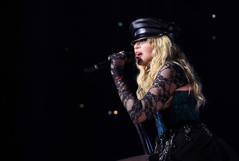 Madonna, wearing a leather biker hat and a laced, low cut black dress with sleeves that cover her hands, sings out to the crowd during a Celebration Tour performance. She has long blonde hair, and the audience is indiscernible amidst the lights.