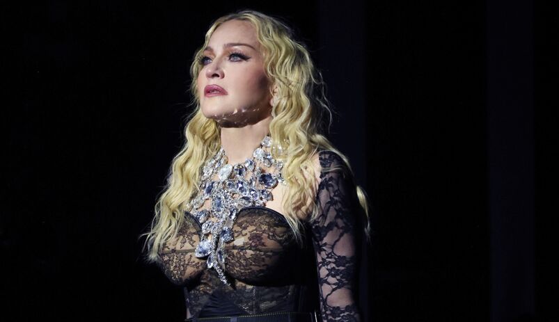 Madonna stands on stage looking off during a performance of her Celebration Tour. She has long blonde hair and wears a large sparkling necklace, draped over a low cut, black laced long sleeved shirt.