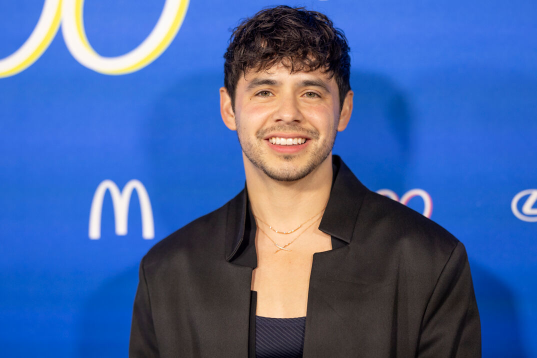 David Archuleta, with short black hair and light dark facial hair, smiles in a low cut navy blue tank top under a black suit jacket. He stands in front of a blue step-and-repeat.