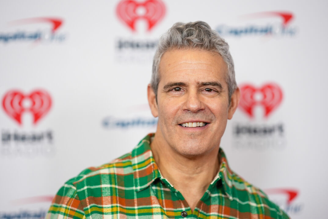 Andy Cohen smiles softly in front of a white step-and-repeat. He has fluffed up gray hair and thin black brows with a clean-shaven face. He wears a green, yellow, and orange pinstriped flannel shirt.