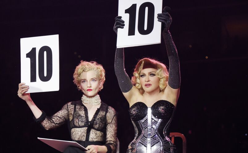 Julia Garner, with short blonde curled hair, pearl necklaces, and a laced black top, holds up a cardboard sign that says "10." Sitting next to her is Madonna, with short blonde hair in a black headband and a tightly fit black and metallic corset. She also holds up the same sign during a Celebration Tour performance.