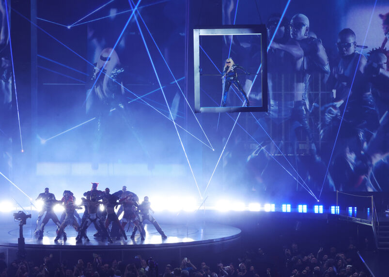 Madonna floats suspended high into the air in a glass box, wearing a tight fitting and sparkling body suit, during a performance of her Celebration Tour. Underneath her on a circular platform, a gang of futuristic dressed dancers pose, while both Madonna and her dancers' likenesses are projected behind them on huge screens.