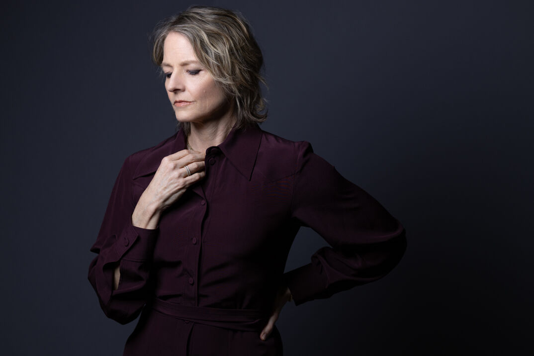 Jodie Foster stands against a gray wall in a longsleeved black blouse. She looks down pensively with her hand to her chest. She has grayed blonde hair down to her shoulder.