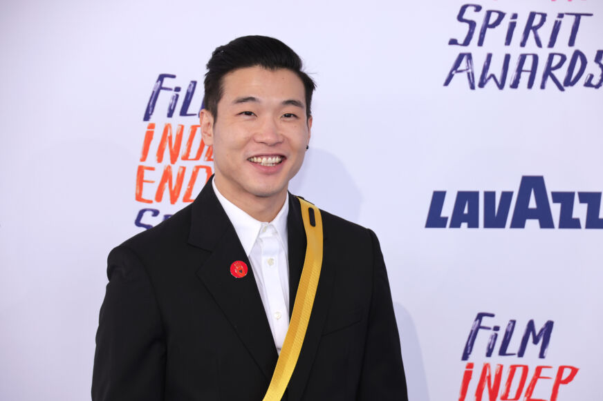Joel Kim Booster smiles in front of a white step-and-repeat wearing a black suit jacket and golden sash over a white dress shirt. He has perfectly coiffed black hair.