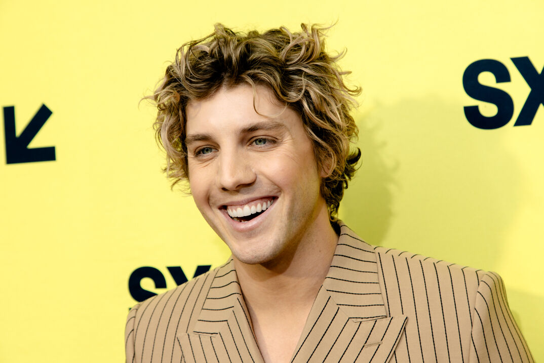 Lukas Gage stands smiling in front of a yellow step-and-repeat at SXSW. He wears a tan suit jacket with thin black lines and has curly dark hair with bleached tips.