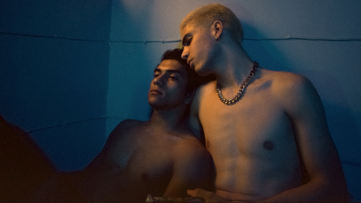 Two shirtless young men—one with bleached hair and a chain necklace—rest on one another against a cement wall.