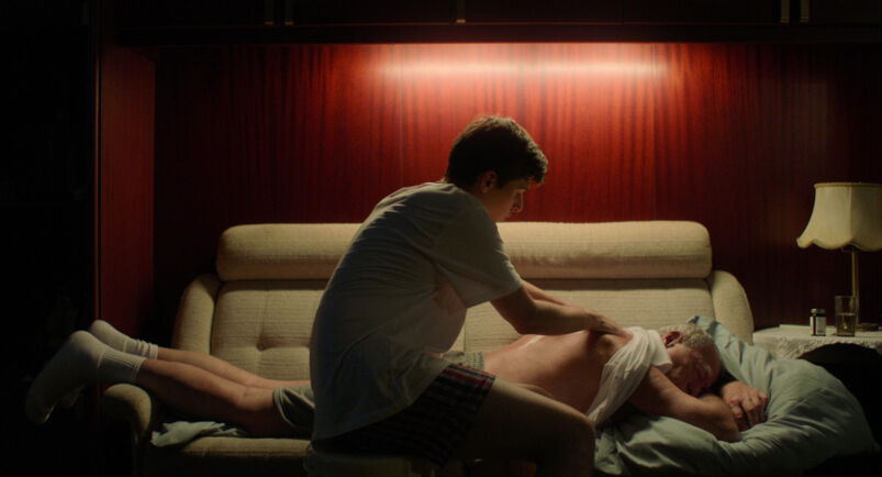A young man in a t-shirt massages the bare back of an older man laying face down on a couch.