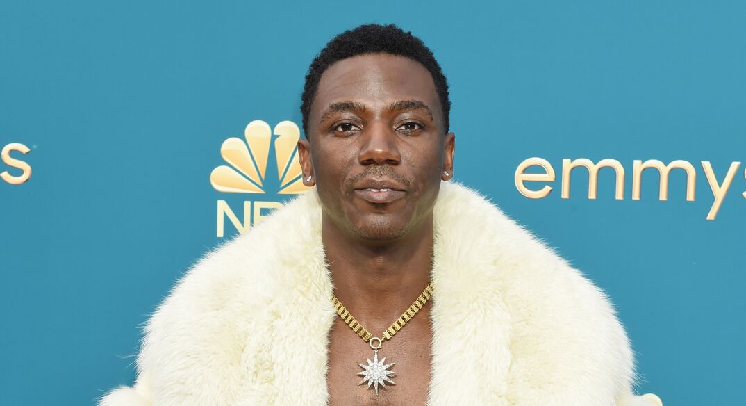 Jerrod Carmichael smiles on the red carpet. He is a Black man with short black hair and a thin mustache. He wears a furry jacket over his bare muscular chest. He wears a diamond star on a gold chain around his neck.