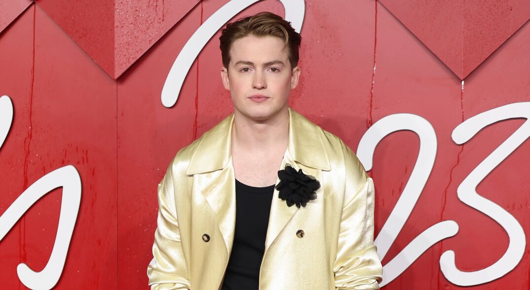 Kit Connor poses at The Fashion Awards 2023 in London England. He has long strawberry blonde hair slicked back and stands in a golden blazer over a blank tanktop.