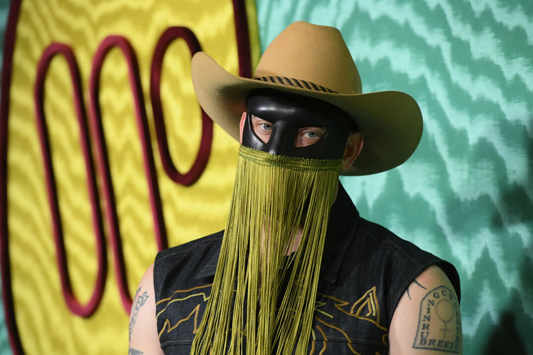 Orville Peck stands on the red carpet, wearing a black mask covering his eyes with long gold fringe that hands past his face. He wears a sleeve black collared shirt and tan cowboy hat. His blue eyes are visible.