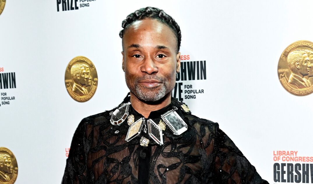 Billy Porter smiles on the red carpet in a black, translucent and sequined dress shirt featuring a collar of large, luxurious diamonds. He has a thin salt and pepper beard and curled locks coiffed on top of his head.